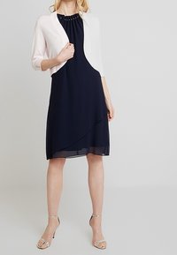 Marinblå klänning med lager-på-lager-kant och pärlbroderad halsringning, kombinerad med en ljusrosa bolero. Slät textur och korta ärmar, silverfärgade sandaler.