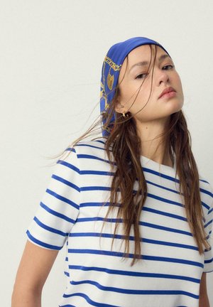 Jonge vrouw met lang bruin haar draagt een wit shirt met blauwe strepen en een blauw patroon hoofddoek, kijkt licht omhoog tegen een eenvoudige achtergrond.