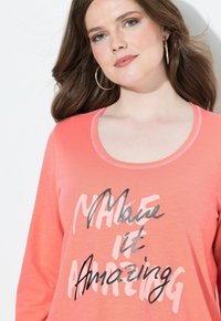 Korallfarbener Langarm-T-Shirt mit einem Rundhalsausschnitt und einem grafischen Druck, auf dem "Make it Amazing" in schwarzer und silberner Schrift steht.