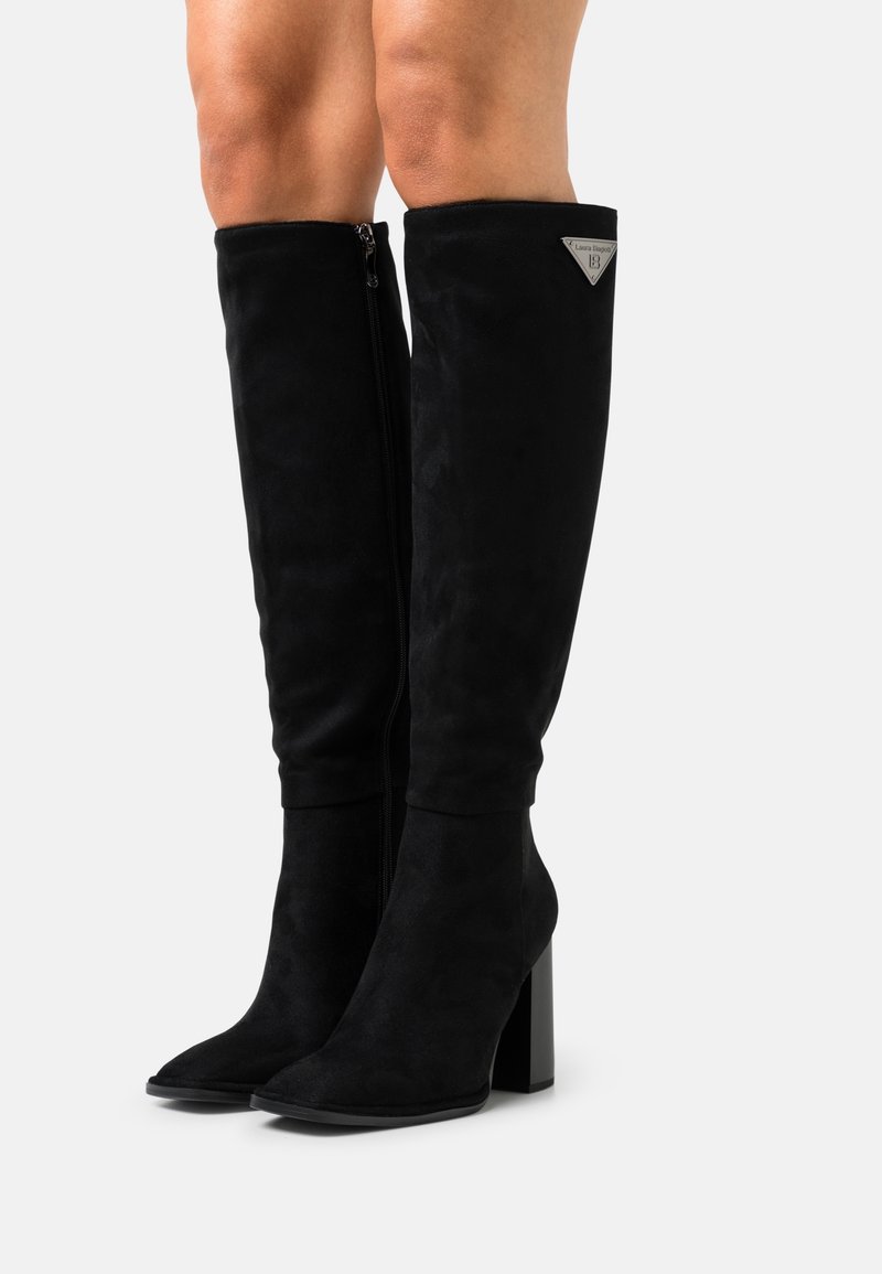Bottes noires en daim jusqu'aux genoux avec des talons carrés et des fermetures éclair latérales portées par une personne avec des jambes nues sur un fond blanc.