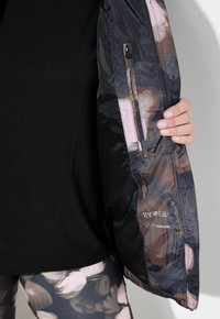 Giacca idrorepellente con esterno lucido e scuro, caratterizzata da un motivo floreale in toni di marrone e rosa tenui, e una tasca interna con zip.