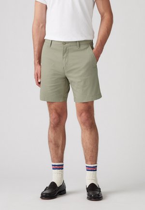 Homme portant un short vert clair, un t-shirt blanc, des chaussettes blanches avec des rayures bleues et rouges, et des mocassins noirs, debout avec une main dans la poche.
