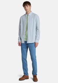 Camisa de botones a rayas en azul, rosa y verde, superpuesta sobre una camiseta verde claro. Combinada con jeans azules y botas de cordones marrones.