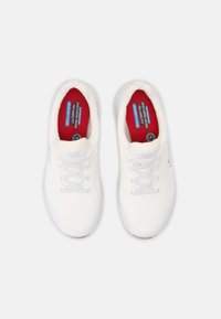 Des baskets blanches antidérapantes avec une tige en tissu texturé, un bout rond, un laçage et des semelles intérieures rembourrées en rouge portant la marque "Mémoire de forme".