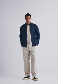 Marineblaues Hemd über einem cremefarbenen Shirt, kombiniert mit beigen Hosen und schwarzen Sneakers mit weißen Schnürsenkeln. Schlichtes, lässiges Design.