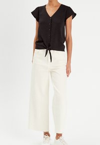 Haut noir à manches courtes avec fermeture à bouton et devant noué, associé à un pantalon large beige clair et des baskets blanches, présenté sur un mannequin.