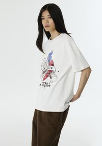 Witte oversized t-shirt met een grafisch ontwerp van een hand die een roos vasthoudt, vlamaccenten en tekst in zwart en rood. Recht gesneden mouwen, relaxte pasvorm.