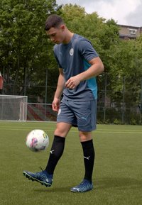 Zili sporta krekli ar īsām piedurknēm, pelēkas shorts, melnas futbolu zeķes un zilas sporta kurpes. Spēlētājs notur multicolor futbolu.