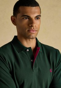 Tume roheline polo särk, millel on magenta krae ja ääred, esiküljel vasakul rinnal väike logo, valmistatud pehmest puuvillast kangast.