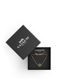 Guldkedjearmband med en grön cirkulär berlock som har en 'C' logotyp, presenterat i en svart texturerad låda med vit branding.