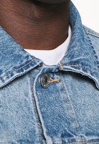 Světle modrý límec džínové bundy s kovovým knoflíkem a šicími detaily. Pod ním je vidět bílý tričko s texturovanou tkaninou.