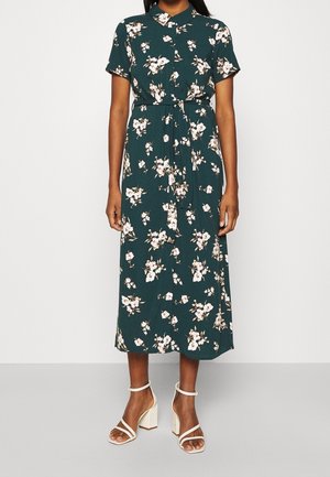 Femme portant une robe midi fleurie boutonnée devant de couleur vert foncé, à manches courtes, avec une ceinture à la taille et des sandales blanches à talons épais.