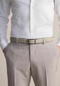 Beige bőr öv sima felülettel és ezüst csattal, világosszürke csíkos öltönynadrág felett és fehér gombos ing alatt viselve.