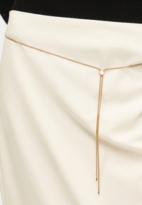 Χρυσή αλυσίδα μέσης με μικρό κυκλικό κούμπωμα και δύο κρεμαστά κεντρικά νήματα φορεμένη πάνω από μια κρεμ φούστα και μαύρο τοπ.