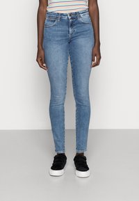 Hellblaue Skinny-Jeans mit mittelhohem Bund, fünf Taschen und kontrastierenden Nähten. Getragen mit schwarzen Sneakers mit strukturiertem Obermaterial.