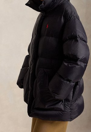 Homme portant une doudoune noire oversize avec un logo rouge brodé sur la poitrine et un pantalon kaki, debout de côté.