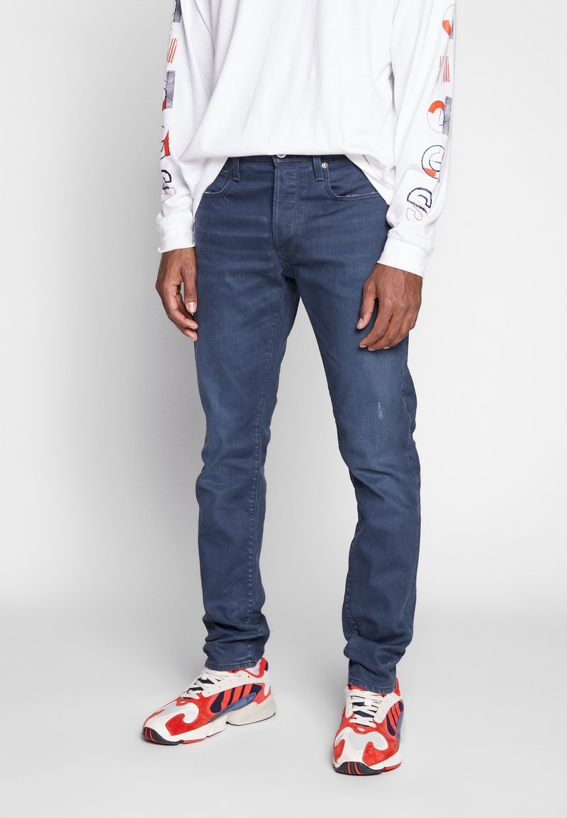 Homme portant un jean slim bleu foncé, une chemise blanche à manches longues avec des graphiques sur les manches, et des baskets blanches avec des accents rouges et bleu marine.