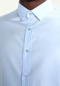 Camisa de botones azul claro con collar clásico, que presenta una textura suave y cinco botones en la parte delantera.