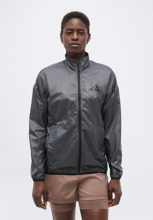 Femeie care poartă o jachetă gri închis cu fermoar și guler înalt și pantaloni scurți bej, stând în fața unui fundal alb simplu.