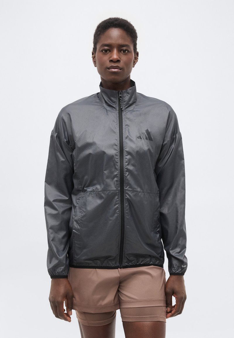 Γυναίκα που φοράει σκούρο γκρι τζάκετ με φερμουάρ και ψηλό γιακά, καθώς και μπεζ σορτς, όρθια μπροστά σε λευκό απλό φόντο.