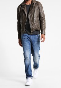 Brun lädermotorcykeljacka med dragkedjor, grå t-shirt, blå denimjeans och vita sneakers, som framhäver en avslappnad, lager-på-lager stil.