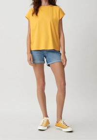 Osoba ubrana w żółtą koszulę z krótkim rękawem, jasnoniebieskie podwinięte dżinsowe szorty i pasujące żółte trampki na białym tle.