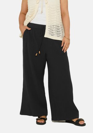 Femme portant un pantalon noir ample à cordon de serrage, un gilet en maille beige par-dessus un haut blanc, et des sandales noires avec une boucle dorée sur un fond clair.