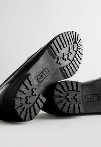 Bottes en caoutchouc noir avec une semelle texturée présentant des crampons prononcés pour une meilleure adhérence. Le mot "NEXT" est embossé sur les deux semelles.