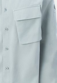 Camisa azul clara de botão feita de tecido suave. Apresenta um bolso no peito e botões padrão com uma textura suave. Design minimalista.