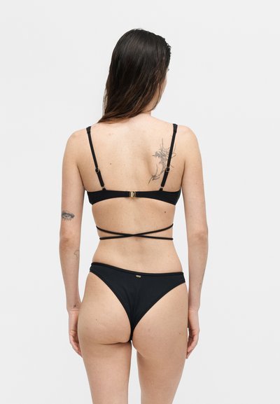 Γυναίκα με μακριά καστανά μαλλιά που φοράει μαύρο μπικίνι με λεπτούς λουριά που σχηματίζουν σταυρό, με τατουάζ στην πάνω πλάτη και στο αριστερό μπράτσο, στέκεται μπροστά σε λευκό φόντο.