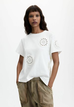 Mujer con cabello oscuro y ondulado que lleva una camiseta blanca de manga corta con patrones de crochet circulares y pantalones marrones sueltos, está de pie con las manos en los bolsillos.