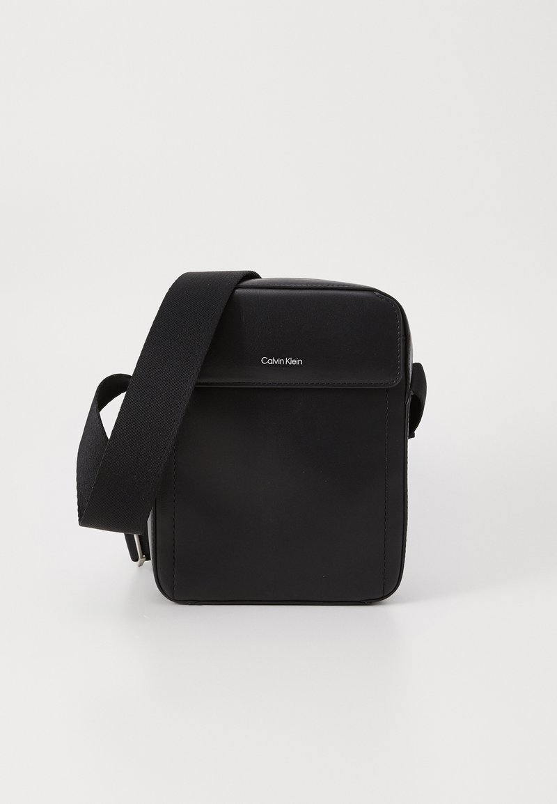 Bolso cruzado de cuero negro con correa ajustable, que presenta una tapa frontal plana, un logo discreto y un diseño rectangular elegante.