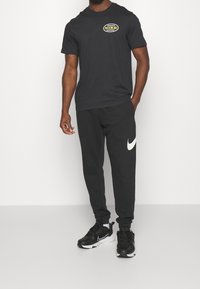 T-shirt en coton noir avec un logo Nike circulaire jaune et vert, associé à un pantalon de survêtement noir avec un swoosh Nike blanc. Baskets noires.