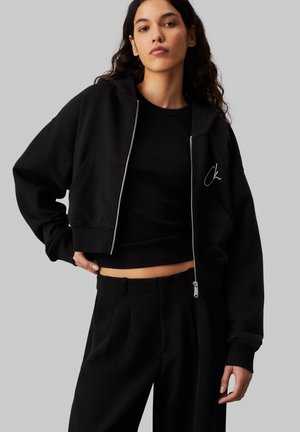 Felpa cropped nera con zip frontale, ricamo con logo e orlo a coste. Abbinata a un top fitted nero e pantaloni sartoriali.