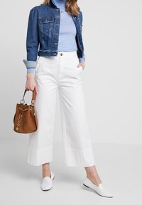 Kvinna i en kort denimjacka, ljusblå polo, vida vita byxor, vita loafers, som håller en brun läderbucketbag med guldkedja.