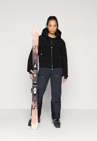 Černá textura bunda, tmavé lyžařské kalhoty, držící růžové a černé lyže. Významné detaily: kapuce, několik zipů a lyžařské vázání.