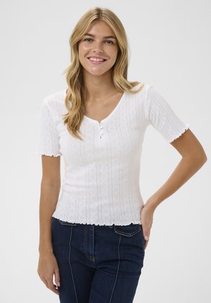 Glunderende vrouw met golvend blond haar draagt een witte, gestructureerde top met korte mouwen en knopen, gecombineerd met donkerblauwe jeans, staand tegen een witte achtergrond.