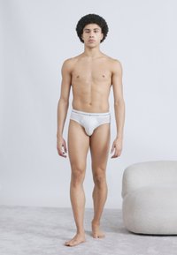 Λευκά ανδρικά εσώρουχα από απαλό ύφασμα, με ελαστική μέση που φέρει λογότυπο. Το μοντέλο στέκεται όρθιο, με φυσική υφή δέρματος σε ουδέτερο φόντο.