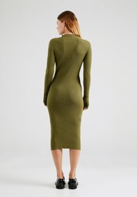 Rippendes olivgrünes Midikleid mit langen Ärmeln, taillierter Silhouette und hohem Kragen. Das Modell kombiniert es mit schwarzen Plateauschuhen.