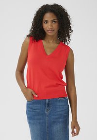 Haut sans manches en tricot rouge avec un décolleté en V, présentant une texture lisse et une coupe décontractée. Associé à une jupe en denim.