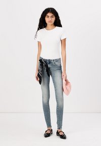 Camiseta blanca de manga corta, jeans ajustados de color azul claro, bandana negra atada a la cintura, bolso de mano rosa y flats negros con hardware decorativo.