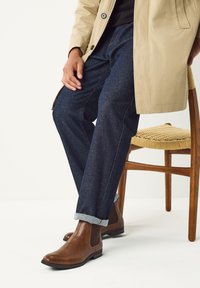 Braune Chelsea-Stiefel aus Leder, dunkelblaue Jeans mit umgekrempelten Säumen und ein beiger Regenmantel, sitzend neben einem Holzstuhl mit geflochtenem Sitz.