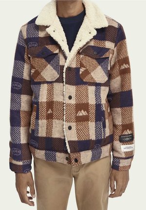 Homme portant une veste à carreaux bleue, marron et beige avec un col en peau lainée et des poches poitrine boutonnées, associée à un pantalon beige.