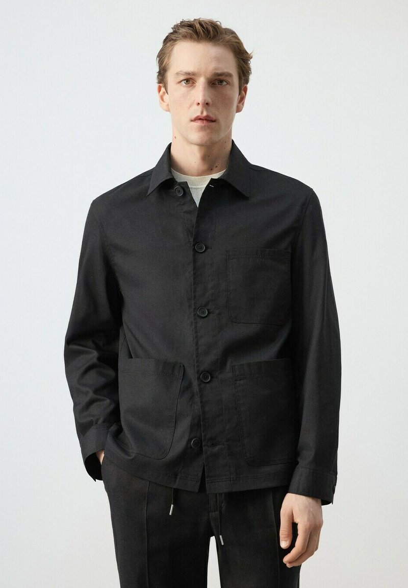 Mladý muž oblečený v čiernej zapínacej bunde s tromi vreckami, stojaci pred jednoduchým bielym pozadím, ruky uvoľnené pozdĺž tela.