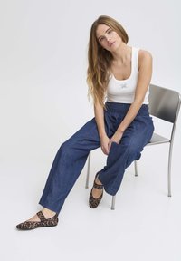 Jeune femme aux cheveux longs assise sur une chaise en métal, portant un débardeur blanc, un pantalon ample bleu marine et des ballerines imprimé léopard.
