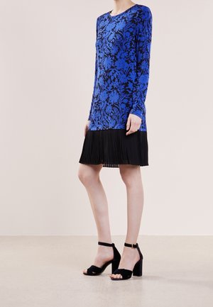 Kobieta ubrana w niebieską, kwiecistą bluzkę z długim rękawem, czarną plisowaną spódnicę oraz czarne sandały na wysokim obcasie z paskiem wokół kostki, stojąca na neutralnym tle.