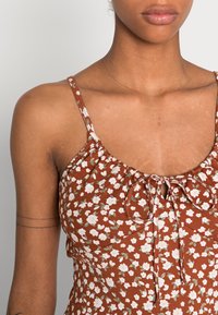 Top floral marrón con tirantes finos, que presenta un escote fruncido y un detalle de lazo en la parte delantera. El material parece suave con una textura ligera.