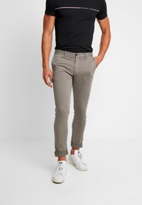 Man som bär khakifärgade upprullade byxor, svart Tommy Hilfiger t-shirt och vita sneakers mot vit bakgrund.