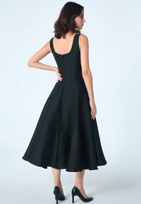 Robe sans manches noire avec un corps ajusté, une jupe évasée et une fermeture éclair dans le dos. Texture lisse et longueur midi. Escarpins noirs portés.