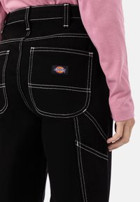 Juodos krovinių kelnės su baltomis siūlėmis, daugybe kišenių ir "Dickies" logotipo etikete. Dėvimos su ilgomis rankovėmis rožine marškinėliais.
